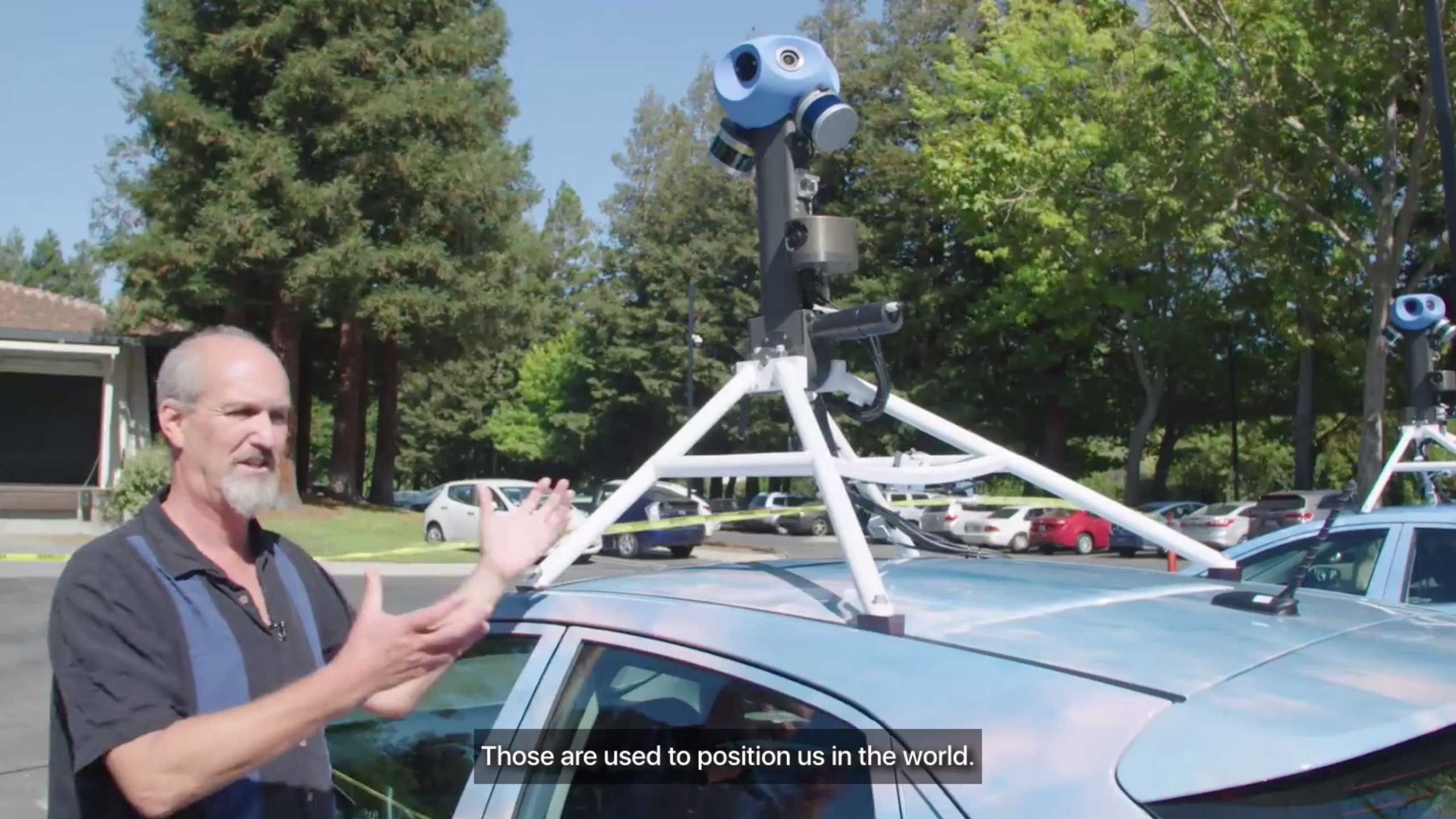 A white man stands beside a car with a camera mounted on its roof. A visible caption says "Those are used to position us in the world."